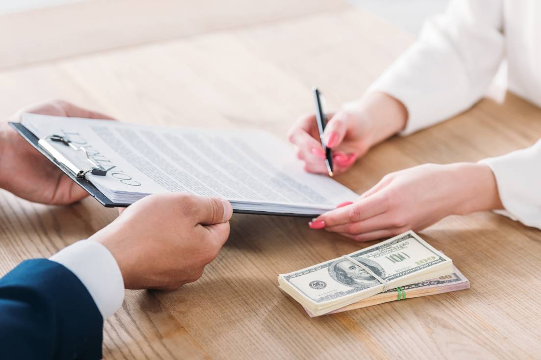 partial view of woman signing contract on clipboard in hands of businessman near dollar banknotes on